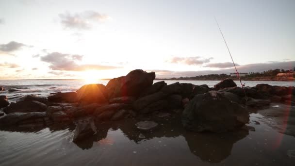 Movimiento lento de la playa a la hora dorada. Primer plano de las rocas en la costa y caña de pescar, mientras que las olas frenan en la arena. Reflejo dorado sobre la superficie del mar. Puesta de sol en el horizonte. Piriapolis, Uruguay — Vídeos de Stock