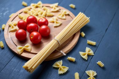 Mavi ahşap arka planda farklı türde çiğ makarna (spagetti, farfalle, fusilli) ile kiraz domatesleri.