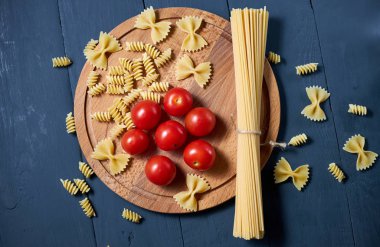 Farklı türde çiğ makarna (spagetti, farfalle, fusilli) ile olgun ve sulu domateslerin üst görünümü.