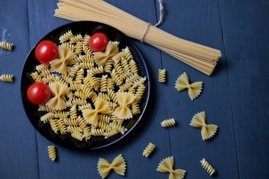 Farklı türde ham makarna (spagetti, farfalle, fusilli) siyah tabakta olgun ve sulu domatesli..