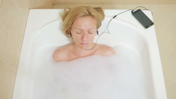 Belle femme prenant un bain et écoutant de la musique sur écouteurs. téléphone portable — Video