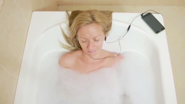 Hermosa mujer tomando un baño y escuchando música en los auriculares. teléfono celular — Vídeo de stock