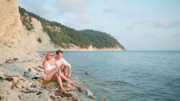 Pareja enamorada está sentada en una roca cerca del mar en unión al atardecer . — Vídeos de Stock