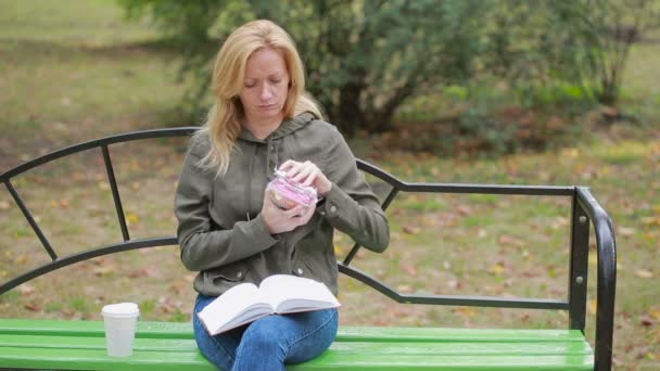 Uma loira num parque a comer um donut. Mulher ao ar livre mastiga pão doce — Vídeo de Stock