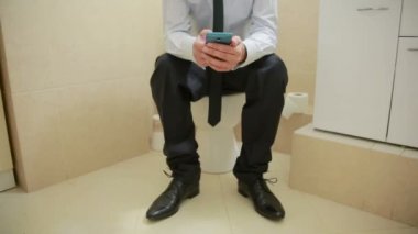 Erkek işadamı tuvalette oturmuş telefonda haberleri izliyor