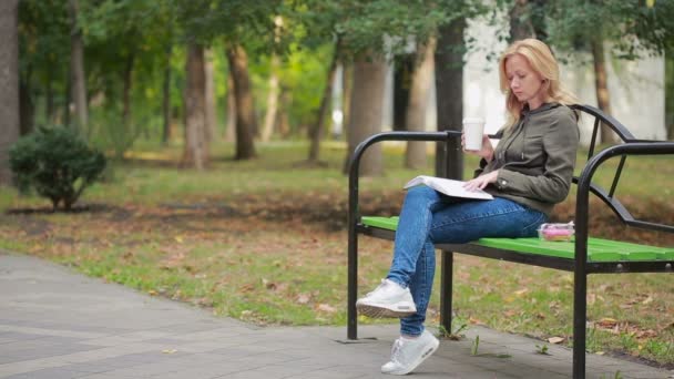 Szőke nő olvasókönyv a fogorvostudomány őszi parkban. forró kávét vagy teát iszik — Stock videók