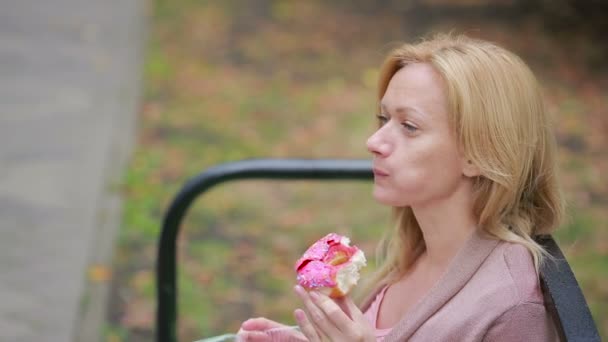 Uma loira num parque a comer um donut. Mulher ao ar livre mastiga pão doce — Vídeo de Stock