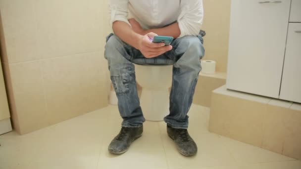 Мужчина сидит на унитазе и смотрит в мобильный телефон — стоковое видео