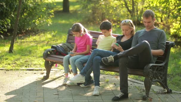 Pais com crianças sentadas no banco e usam smartphones móveis — Vídeo de Stock