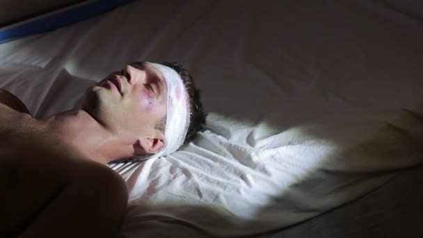 Mężczyzna z obrażeniami głowy, leżąc na łóżku. obandażowaną głową. siniaki na twarzy. — Wideo stockowe