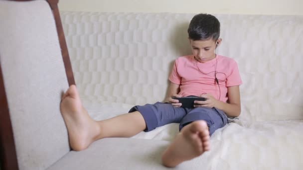 Αγόρι με τα smartphone και τα ακουστικά να ακούτε μουσική ή να παίζει το παιχνίδι στο σπίτι — Αρχείο Βίντεο