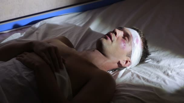Un hombre con lesiones en la cabeza acostado en la cama. cabeza vendada. moretones en la cara . — Vídeo de stock