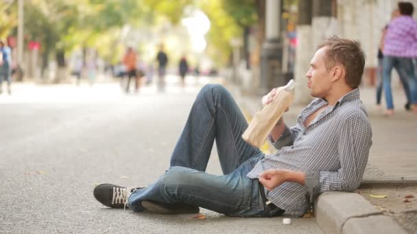 П'яний чоловік лежить на тротуарі зовні. чоловік п'є пиво з паперового мішка — стокове відео