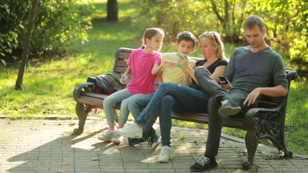 Rodiče s dětmi, sedí na lavičce a využití mobilní smartphony — Stock video