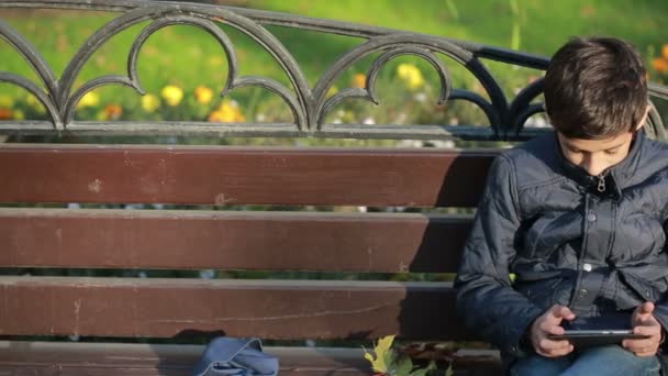 Αγόρι σε εξωτερικούς χώρους το φθινόπωρο που κάθονται στον πάγκο σε ένα σακάκι και χρησιμοποιήστε ένα smartphone — Αρχείο Βίντεο