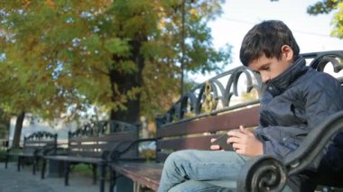 Çocuk açık havada bir ceket ve kullanımı bir smartphone bankta oturmuş Güz