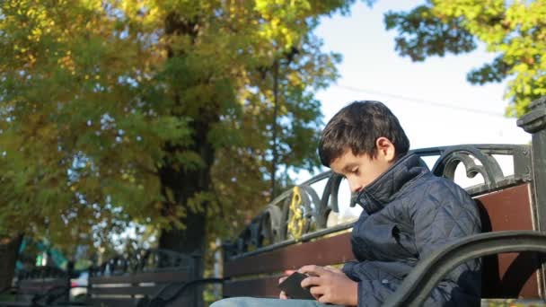 Niño al aire libre en otoño sentado en el banco en una chaqueta y utilizar un teléfono inteligente — Vídeos de Stock