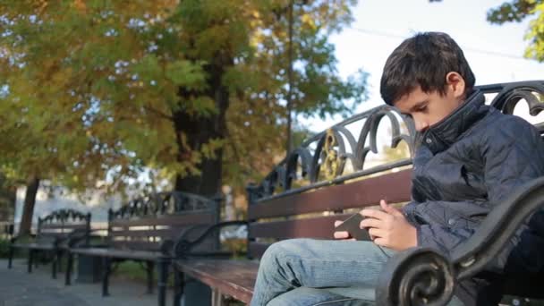 Niño al aire libre en otoño sentado en el banco en una chaqueta y utilizar un teléfono inteligente — Vídeos de Stock