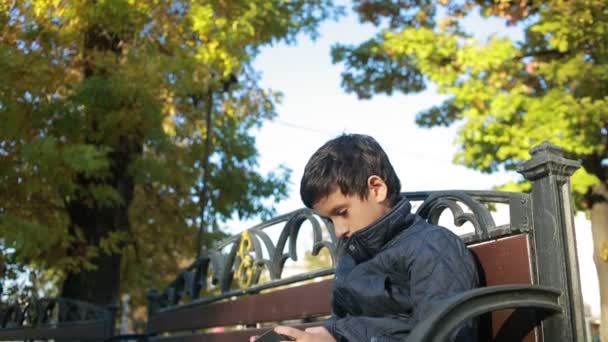 Niño al aire libre en otoño sentado en el banco en una chaqueta y utilizar un teléfono inteligente — Vídeo de stock