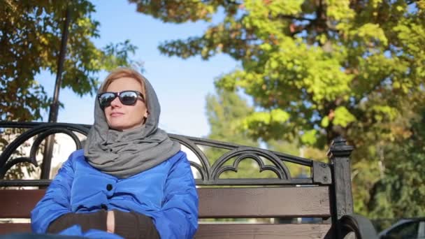 Vakker kvinne i solbriller som sitter på benken i høstparken. Gule blader – stockvideo