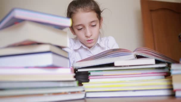 Liten flicka i skoluniform läser en bok, sitter mellan högar av böcker — Stockvideo
