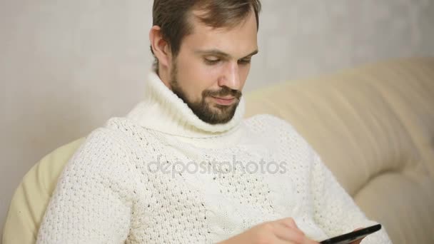 Jovem barbudo homem em uma jaqueta no sofá com um tablet — Vídeo de Stock