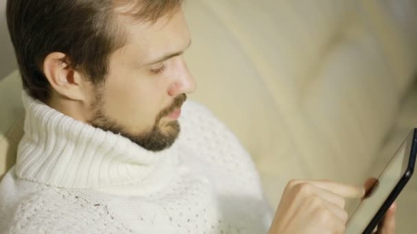 Молодий бородатий чоловік в куртці на дивані з таблеткою — стокове відео