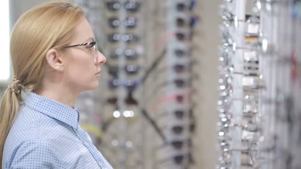 Женщина блондинка выбирает новые очки в оптике — стоковое видео