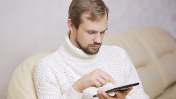 Młody, brodaty mężczyzna w kurtkę na kanapie z tabletem — Wideo stockowe
