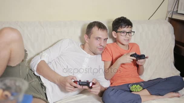 Отец и сын играют в видеоигры в своем доме на диване — стоковое видео