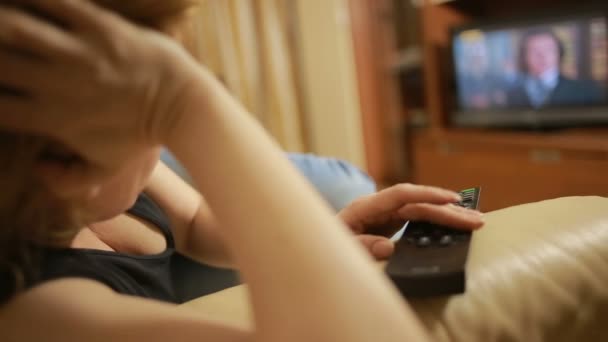 Красивая женщина смотрит телевизор сидя дома на диване, переключает каналы — стоковое видео