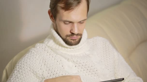 Young bearded man in a jacket on the couch with a tablet — Stock Video