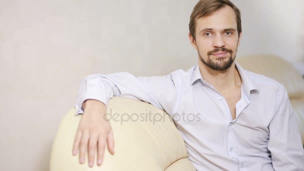 Un portraitiste regardant la caméra sur le canapé. Jeune homme avec chemise relaxante à la maison — Video