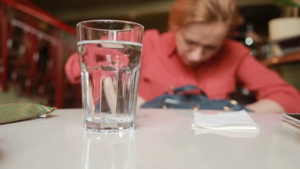 Fată în cafenea este în căutarea pentru ceva într-o pungă pe un fundal de sticlă cu apă — Videoclip de stoc