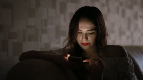 Femme avec un téléphone dans le noir à la maison sur le canapé. téléphone reflet visage — Video