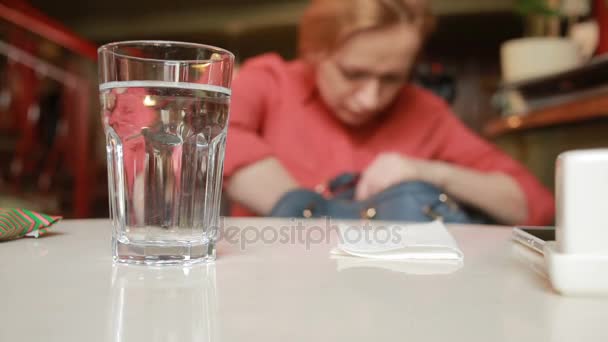 カフェで女の子が水でガラスの背景に袋に何かを探しています。 — ストック動画