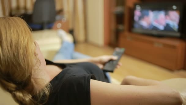 Όμορφη γυναίκα βλέποντας τηλεόραση καθισμένος στον καναπέ στο σπίτι, αλλάζει κανάλια — Αρχείο Βίντεο