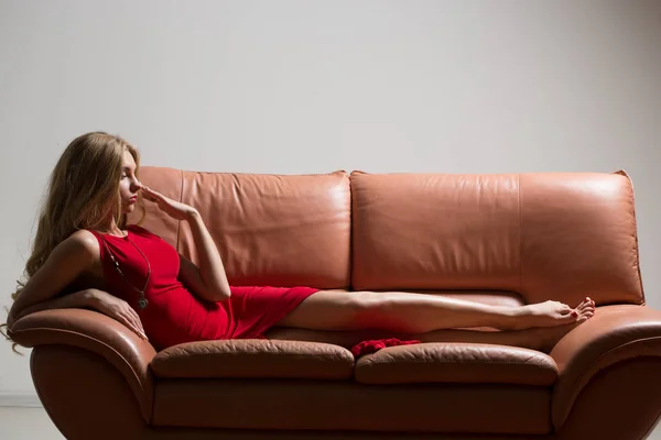 Ritratto a figura intera di bella giovane donna bionda sul divano. lunghi capelli ricci. Abito rosso — Foto Stock