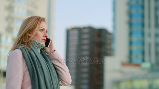 Meisje in een roze jas met telefoon loopt op een moderne stad. achtergrond van wolkenkrabbers — Stockvideo