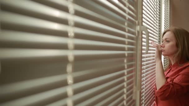 Mujer mirando por la ventana a través de las persianas a la calle, espiando. Sospechada — Vídeos de Stock