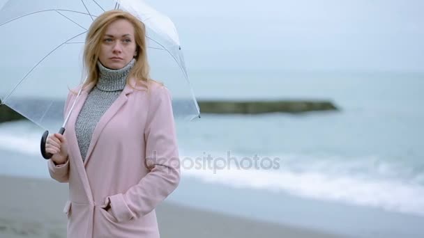 Девушка в розовом пальто с прозрачным зонтиком у моря во время шторма . — стоковое видео