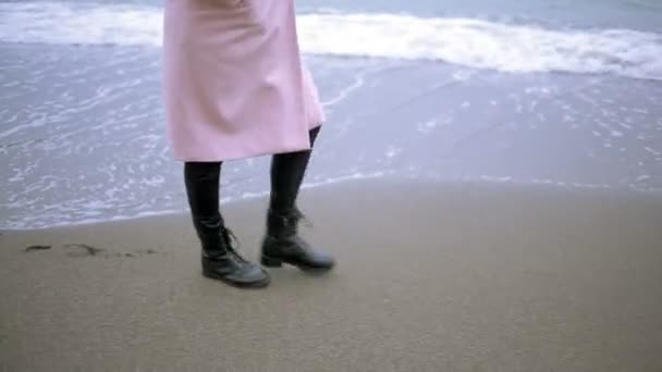 Счастливая девушка в розовом пальто у моря во время шторма. Море смывает свои следы в песке — стоковое видео