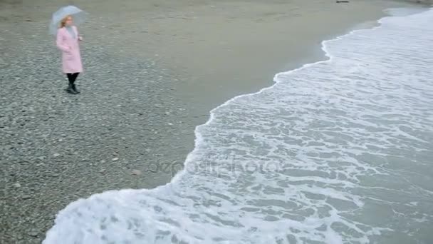 Menina em um casaco rosa com um guarda-chuva transparente junto ao mar durante uma tempestade . — Vídeo de Stock