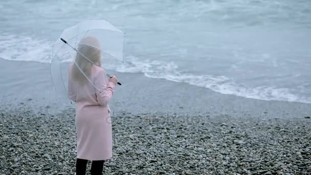Ragazza in cappotto rosa con un ombrello trasparente in riva al mare durante una tempesta . — Video Stock