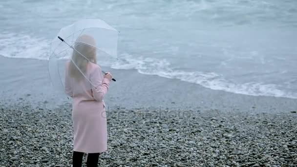 Niña en un abrigo rosa con un paraguas transparente junto al mar durante una tormenta . — Vídeos de Stock