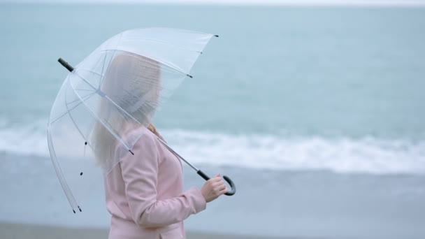 Fille en manteau rose avec un parapluie transparent au bord de la mer pendant une tempête . — Video