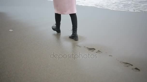 Szczęśliwa dziewczyna w różowy płaszcz, w pobliżu morza podczas burzy. Morze myje jej ślady stóp na piasku — Wideo stockowe