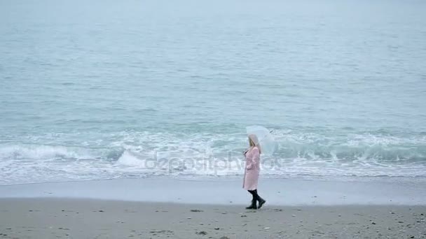Niña en un abrigo rosa con un paraguas transparente junto al mar durante una tormenta . — Vídeo de stock