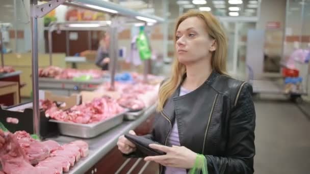 Mujer en un supermercado junto a un mostrador de carne, comprando carne, mira a la cámara — Vídeos de Stock