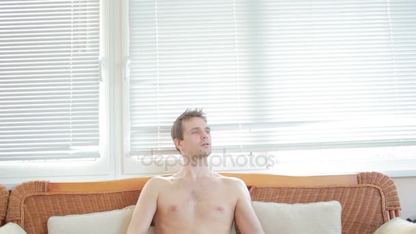 Un hombre con un torso desnudo se sienta en un sofá de mimbre y toca la guitarra — Vídeo de stock
